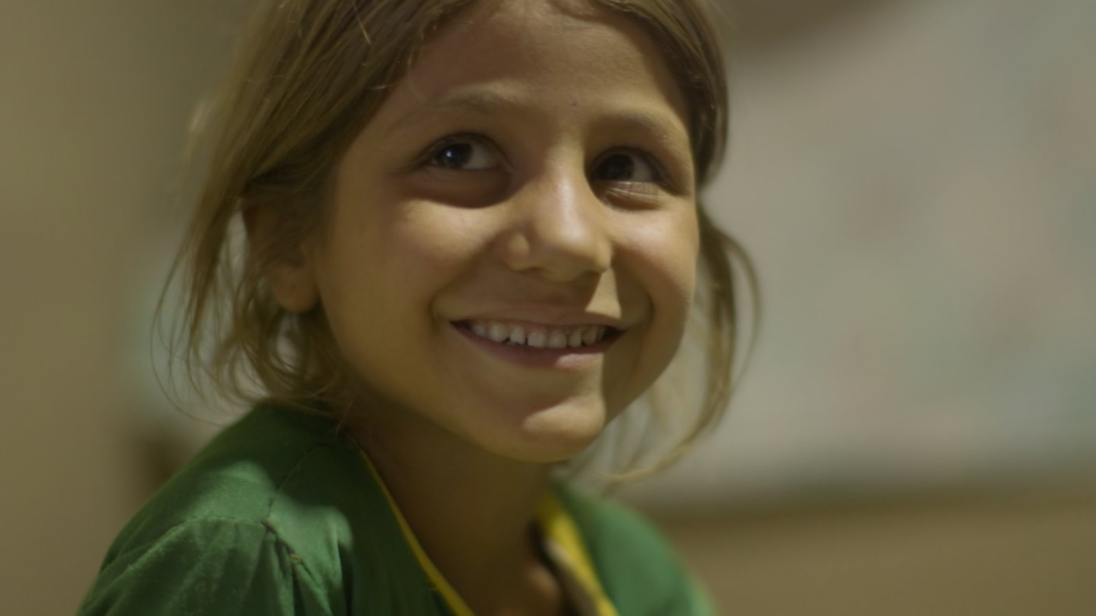 A young Afghan girl in a green dress smiling warmly, photographed in Herat