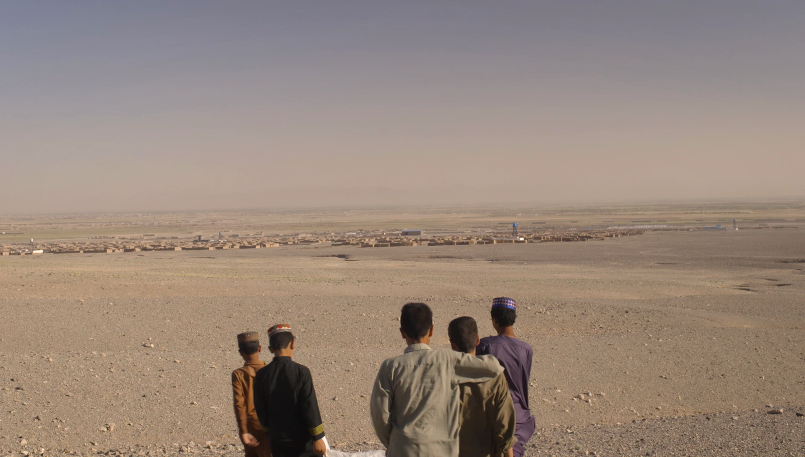 Four Afghan children standing together looking out over a vast desert landscape toward a distant town, with open sky above